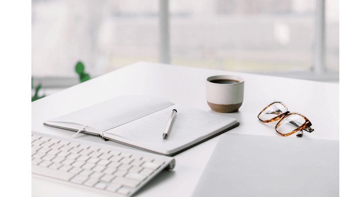 white desk with keyboard, notepad, pen, glasses, and coffee mug