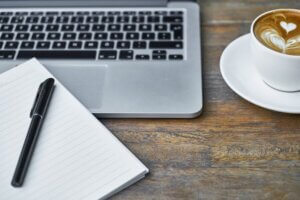 desk with laptop, coffee cup with coffee and saucer, and notebook with black pen