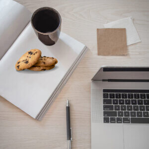 laptop on desk with open blank book. Mug and chocolate chip cookies sitting on top of it